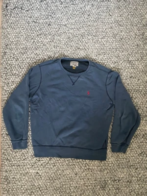 Blå crewneck från Polo Ralph Lauren - Snygg blå crewneck från Polo Ralph Lauren med ganska stor klassisk rund halsringning, perfekt för en skjorta eller en polo under, och broderad röd logga på bröstet. Tröjan har ribbade muddar vid ärmslut och nederkant. Perfekt för en avslappnad och stilren look. Strl L herr, passform kolla bild, för referens 184
