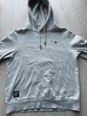 Ljusgrå hoodie från Morris - Ljusgrå hoodie från Morris med broderad svart lilja på bröstet och patch nertill. Klassisk modell med dragsko i huvan och ribbade muddar. Perfekt för en avslappnad och stilren look. Är lite liten i storleken men ska passa alla från 155-165cm