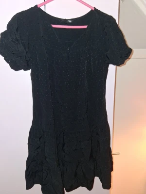 Svart kortärmad klänning H&M - Snygg svart klänning från H&M med korta puffärmar och diskret prickigt mönster. Klänningen har v-ringning med en liten knapp framtill och är i en kort modell. Perfekt för dig som gillar enkel men stilren design.