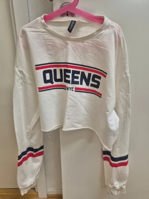 Vit croppad långärmad topp Divided S - Säljer en vit croppad långärmad topp från Divided H&M med trycket 'QUEENS NYC' i blått och rött framtill. 