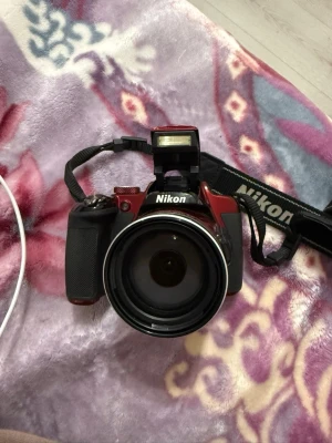 Röd Nikon digitalkamera P610 - Säljer en röd Nikon Coolpix P610 digitalkamera med utfällbar blixt och vridbar skärm. Kameran har många knappar och reglage för enkel kontroll, samt ett bekvämt grepp och en svart axelrem. Perfekt för dig som vill ta snygga bilder och filma.