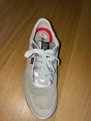 Beige sneakers från Callaghan - Snygga beige sneakers från Callaghan med mesh- och mockadetaljer. Skorna har vita snören, en diskret röd logga på sidan och silvriga detaljer vid hälen. Insidan har en röd kant och sulan är vit och rejäl för extra komfort.