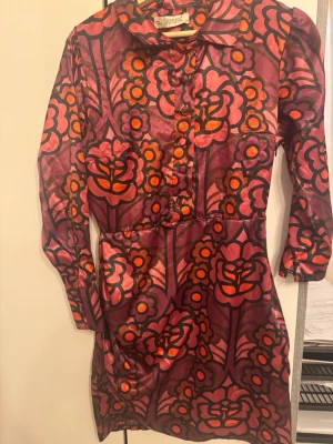 Blommig lila klänning från Şamdan - Färgstark klänning från Şamdan med blommigt mönster i lila, rosa och orange toner. Klänningen har långa ärmar, krage och knappar framtill. Sidenliknande material ger en glansig finish och retrovibbar.