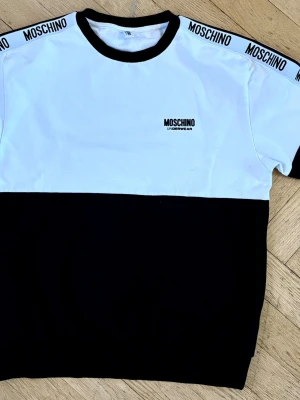 Svartvit tröja från Moschino - Snygg svartvit tröja från Moschino med logga på bröstet och textdetaljer på axlarna/ärmarna. Perfekt för dig som gillar streetwear och vill sticka ut med ett trendigt plagg.