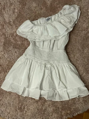 Vit kortklänning med volang - Supersöt vit kortklänning från MEET ME THERE med bred volang upptill och smock i midjan. Aldrig använd