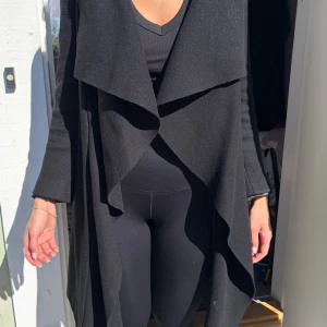 Svart lång kappa med vattenfallsärm,S-M - Stilren svart kappa med vattenfallsinspirerad krage och öppen front. Kappan har lång ärm och en lätt utsvängd passform baktill. Perfekt för dig som gillar en klassisk men modern look. Materialet känns mjukt och följsamt.