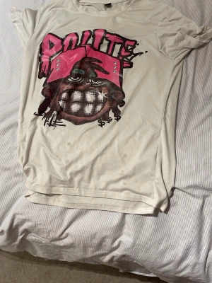 Vit t-shirt med grafiskt tryck - Säljer en vit t-shirt med ett stort grafiskt tryck framtill i rosa och svart. Trycket har en cool tecknad figur med bling och texten 'POLITE' i graffiti-stil. T-shirten har rund hals och korta ärmar, perfekt för en avslappnad streetwear-look.