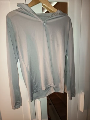 Ljusgrå träningströja med dragkedja - Ljusgrå träningströja i tunt och stretchigt funktionsmaterial. Tröjan har lång ärm, huva och en halv dragkedja framtill. Enkel och stilren design utan synliga loggor eller mönster, perfekt för träning eller chill.