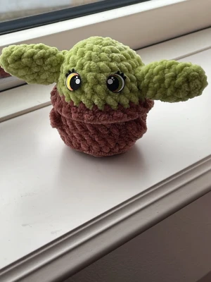 Virkad Baby Yoda mjukisfigur - Söt virkad Baby Yoda-inspirerad mjukisfigur med stora gröna öron och brun kropp. Tillverkad i mjukt, fluffigt garn och har bruna/guldfärgade filtögon med vita detaljer. Perfekt som studentpresent eller present till Star Wars-fans.