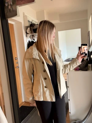 Trenchcoat  - Kort Beige trenchcoat men ett band rund midjan man kan justera, både snyggt utan som med band. Storlek S