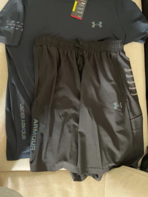 Under Armour Tracksuit - Snyggt svart träningsset från Under Armour med t-shirt och shorts. T-shirten har logga på bröstet och ärmen, medan shortsen har elastisk midja och diskret logga på benet. Perfekt för gymmet eller löprundan. Materialet är lätt och andas bra.