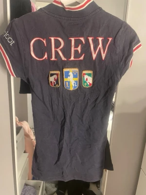 Blå Falsterbo piké från Gina Tricot - Mörkblå pikétröja från Gina Tricot med broderade märken och texten 'CREW' på ryggen. Framsidan har Falsterbo Horse Show-brodyr och knappar vid halsen. Rödvita detaljer på krage och ärmslut ger en sportig look.
