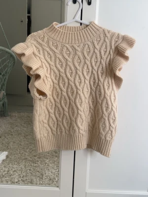 Beige stickad väst med volangärm - Supersöt beige stickad väst med volangdetaljer vid ärmarna och ett snyggt kabelstickat mönster. Perfekt att styla över en skjorta eller t-shirt för en trendig look. Ribbad kant vid hals, ärm och nederkant.
