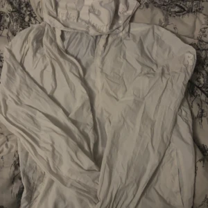 Risky vindjacka vit - Säljer en stilren vit vindjacka med dragkedja och huva. Jackan har en enkel design och är tillverkad i ett lätt material som passar perfekt för blåsiga dagar. Resår vid ärmslut och nederkant ger en skön passform.