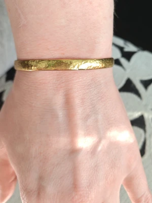 Hammrat guldigt armband i metall - Snyggt armband i guldig metall med hamrad yta som ger en cool och unik look. Armbandet är stelt och öppet i bak, vilket gör det lätt att ta på och av. Perfekt accessoar för att lyfta vilken outfit som helst.