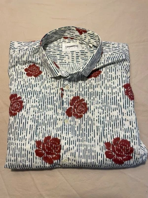 Köp skjortan☺️ - Säljer en vit skjorta från Lindbergh i storlek M med röda blommor och tunna svarta ränder. Skjortan har klassisk krage, knappar framtill och långa ärmar. Köp den ba🥰