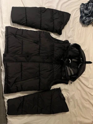 Svart dunjacka från Burberry - Snygg svart dunjacka från Burberry med avtagbara ärmar. Jackan har dragkedjor, praktiska fickor och en tydlig Burberry London England-logga på ärmen. Perfekt för lager-på-lager och streetstyle. Materialet är klassiskt dun för extra värme.