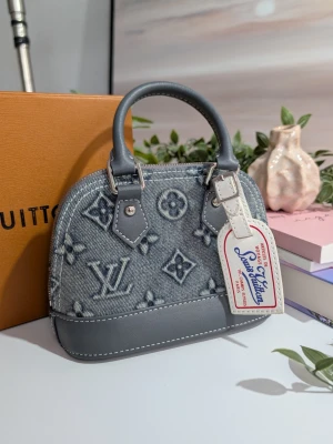 Iouis Vuitton handväska i miniatyr - En handväska i miniatyr med rundad ovansida, tillverkad i blå denim med ett ljusgrått mönster av blommor och symboler. Den har detaljer i gråblått läder, vita kontrastsömmar och korta runda handtag. På framsidan hänger en vit dekorativ etikett med röd och blå text.