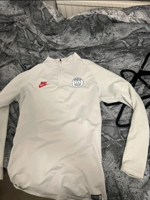 Vit Nike PSG träningströja - Vit långärmad träningströja från Nike med PSG-logga på bröstet och röd Nike-symbol. Tröjan har halv dragkedja framtill och är tillverkad i ett lätt material som passar för sport och aktivitet.