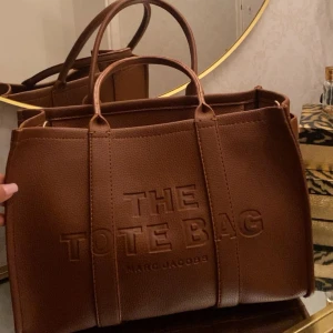 Brun Tote Bag  - Säljer en snygg brun Tote Bag från Marc Jacobs i strukturerat skinn. Väskan har präglad logga framtill, dubbla handtag och rymligt fack med innerficka och dragkedja. Perfekt för dig som vill ha en stilren och trendig väska med plats för allt du behöver.