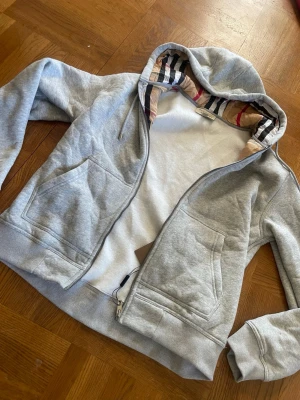 Grå Burberry zip hoodie - Snygg grå hoodie från Burberry med dragkedja och klassiskt rutigt foder i huvan. Hoodien har två stora fickor framtill och ribbade muddar. Perfekt för dig som gillar stilrena plagg med exklusiva detaljer.                                                          Skickar inom 24h✅