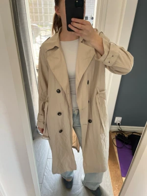 Beige trenchcoat  - Stilren lång beige trenchcoat med dubbelknäppning och två stora fickor framtill. Som referens är jag 170cm lång 