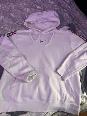 Vit hoodie från Nike med logga - Snygg vit hoodie från Nike med svart broderad logga på bröstet och färgade detaljer på axlarna. Klassisk känguruficka framtill och dragsko i huvan. Mjuk insida och ribbade muddar vid ärmslut och nederkant. Perfekt för en sportig och avslappnad stil.