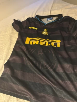 Inter Milan Umbro fotbollströja Ronaldo - Säljer en svart och mörkblå randig Inter Milan fotbollströja från Umbro med gula detaljer, Pirelli-logga och klubbmärke framtill. På ryggen står det Ronaldo och nummer 10 i gult. Tröjan har korta ärmar och är i lätt, ventilerande material.