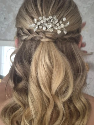 Hårkam med glittrande stenar - Snygg hårkam i silverfärgad metall dekorerad med klara, glittrande stenar i olika former. Perfekt för att skapa en elegant look med extra bling. Kammen har en blomliknande design och passar till uppsättningar eller lockigt hår. Använd 1 gång och sitter bra hela kvällen.