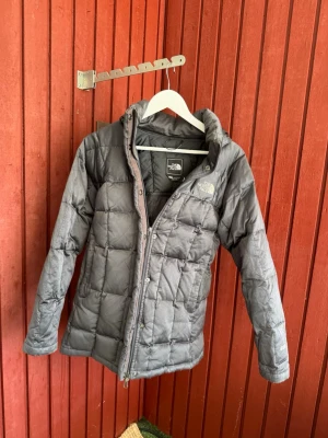 Grå dunjacka från The North Face - Snygg grå dunjacka från The North Face med quiltat mönster och huva. Jackan har dragkedja och tryckknappar framtill samt logga på bröstet. Perfekt för kalla vinterdagar och håller dig varm med sin dunfyllning.