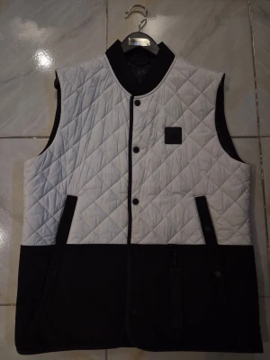 Vit och svart dunväst Moose Knuckles - Snygg quiltad dunväst från Moose Knuckles i vitt och svart. Västen har svarta detaljer, knappar framtill och två fickor. Märkets logga sitter på bröstet. Perfekt för lager-på-lager och streetstyle.