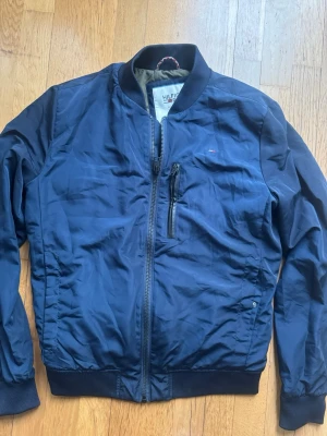 Snygg marinblå bomberjacka Tommy Hilfiger - Snygg marinblå bomberjacka från Tommy Hilfiger med dragkedja framtill, ribbade muddar och en bröstficka med dragkedja. Klassisk bombermodell med liten logga på bröstet och mjukt innerfoder. Perfekt för dig som gillar stilren och sportig look.
