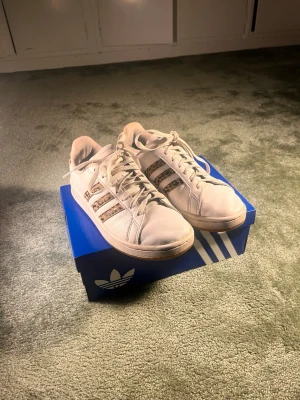 Vita Adidas sneakers med leoparddetaljer - Säljer ett par vita Adidas sneakers med klassiska tre ränder på sidan, där ränderna har coola leopardmönstrade detaljer. Skorna har snörning och guldlogga på tungan. Perfekta för dig som vill sticka ut med en unik touch på dina sneakers.