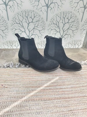 Svarta Chelsea boots i mocka - Säljer ett par klassiska svarta Chelsea boots i mocka med elastiska sidpaneler och dragflik bak. Bootsens stilrena design gör dem enkla att matcha och de har en låg klack och rundad tå. Perfekta för dig som gillar tidlösa och snygga skor.