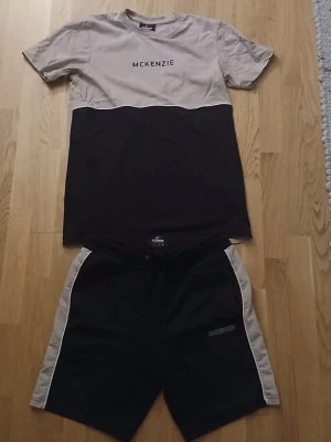 Svart och beige set från McKenzie - Tvådelat set från McKenzie med en t-shirt och shorts. T-shirten är beige upptill och svart nedtill med McKenzie-logga på bröstet. Shortsen är svarta med beige sidor och har en liten logga på vänster ben. Perfekt för en avslappnad stil.