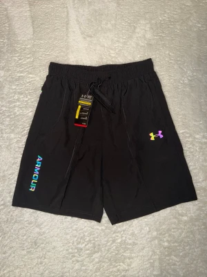 Under armour reflective shorts - Säljer ett par svarta Under Armour shorts med reflex logga och text på vänster ben. Shortsen har elastisk midja med snörning och är tillverkade i ett lätt och snabbtorkande material. Perfekta för gymmet eller löprundan.