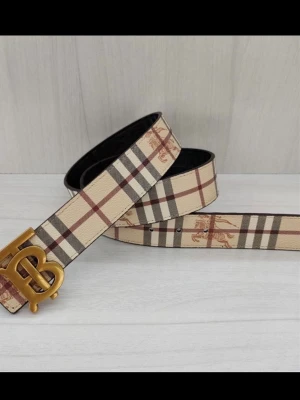 Beige rutigt Burberry bälte - Snyggt bälte från Burberry i klassiskt beige rutigt mönster med guldigt BB-spänne. Bältet har detaljer i brunt, svart och rött och är gjort i skinn. Perfekt accessoar för att lyfta vilken outfit som helst.