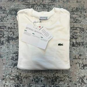 Vit t-shirt från Lacoste - Klassisk vit t-shirt från Lacoste med rund halsringning och den ikoniska gröna krokodilloggan broderad på bröstet. Tillverkad i mjuk bomull för en skön känsla och enkel stil. Perfekt basplagg för en clean och stilren look.