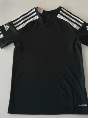 Svart Adidas t-shirt med vita ränder - Svart t-shirt från Adidas med klassiska vita ränder på axlarna och ärmarna. Adidas-logga på vänster ärm och Aeroready-tryck nedtill. T-shirten har rund hals och är tillverkad i ett lätt och sportigt material.