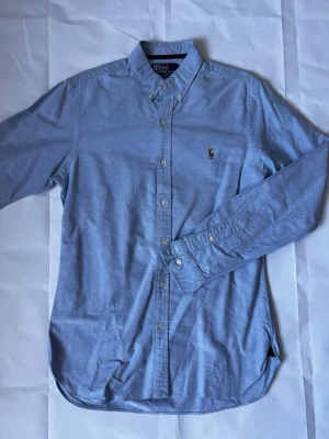 Blå skjorta från Polo Ralph Lauren - Klassisk ljusblå skjorta från Polo Ralph Lauren med button-down krage och broderad logga på bröstet. Skjortan har långa ärmar och knäppning framtill. Perfekt för en stilren och avslappnad look.
