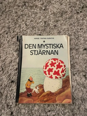 Den mystiska stjärnan Tintins äventyr - Säljer seriealbumet 'Den mystiska stjärnan' från Tintins äventyr av Hergé. Omslaget är vitt med färgglad illustration av Tintin och Milou vid en stor svamp. Boken är i mjukpärm och har klassisk tecknad stil.