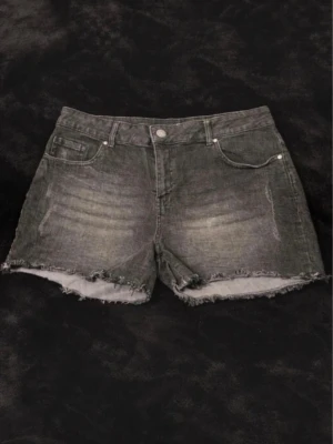 Grå jeansshorts med fransig kant - Snygga grå jeansshorts med fransig, rå kant och klassisk femficksdesign. Shortsen har en något tvättad look och stängs med knapp och dragkedja. Perfekta för en avslappnad stil under varmare dagar.
