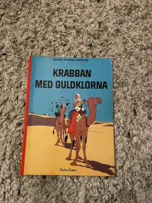Krabban med guldklorna Tintin - Säljer seriealbumet 'Krabban med guldklorna' från Hergés klassiska Tintin-serie. Omslaget är blått och gult med illustration av Tintin och kapten Haddock på kameler i öknen. Boken är i hårdpärm och har färgglada teckningar genom hela albumet.