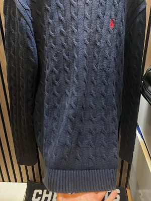 Mörkblå kabelstickad tröja Polo Ralph Lauren - Kabelstickad mörkblå tröja från Polo Ralph Lauren med klassisk röd logga broderad på bröstet. Tröjan har rund halsringning, långa ärmar och ribbade muddar vid ärmslut och nederkant. Perfekt för en stilren och avslappnad look.