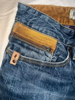 Blå jeans med senapsgula detaljer - Säljer ett par blå regular tapered jeans från Denim & Co med unika senapsgula manchesterdetaljer i linningen och på fickan. Jeansen har dragkedja på fickan och snygga kontrastsömmar. Perfekta för dig som vill sticka ut lite extra!