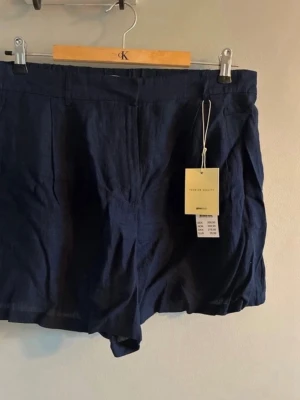 Mörkblå linneshorts från Gina Tricot - Snygga mörkblå shorts från Gina Tricot i luftigt linne. Klassisk modell med hög midja och diskreta fickor i sidorna. Perfekta för varma dagar och enkel att matcha med olika toppar.