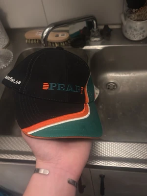 Kneg keps - Snygg svart keps från peab  med broderad logga framtill i turkos och orange. Skärmen har coola detaljer i grönt, orange och vitt. Klassisk form med böjd skärm och justerbar baksida. Perfekt för att lyfta din streetwear-look. Kamma hem han för 4500, pris kan diskuteras