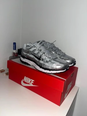 Nike silverfärgade sneakers - Snygga sneakers från Nike i metallic silver med svarta och vita detaljer. Skorna har en sportig design med mesh och syntetmaterial, samt en svart swoosh på sidan. Sulan är vit med svarta inslag och ger en modern look. Perfekta för dig som gillar streetwear.