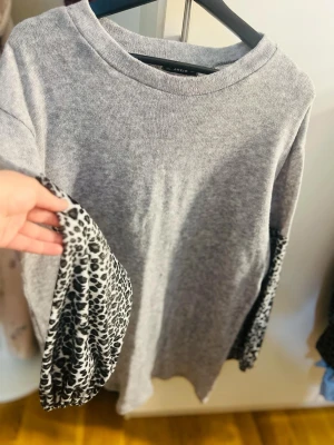 Grå tröja med leopardmönstrade ärmar - Cool grå tröja från Gina Tricot med unika leopardmönstrade ärmar i svart och vitt. Tröjan har rund halsringning och en avslappnad passform. Perfekt för dig som vill sticka ut med en trendig detalj.