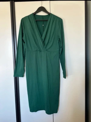 Grön klänning med lång ärm - Stilren grön klänning med omlott-v-ringning och långa ärmar. Modellen är knälång och har en enkel, elegant design som passar perfekt till olika tillfällen. Tillverkad i mjukt material för en bekväm känsla.